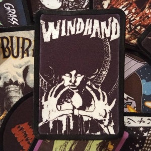 May include: Black and white embroidered patch with the text "WINDHAND" in white lettering. The patch features a stylized image of a person with their hands raised in a gesture of worship.