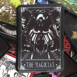 Peut inclure: Patch brodé en noir et blanc avec une figure à capuche tenant un bâton et une coupe. Le patch a une bordure noire et le texte "THE MAGICIAN" en bas.