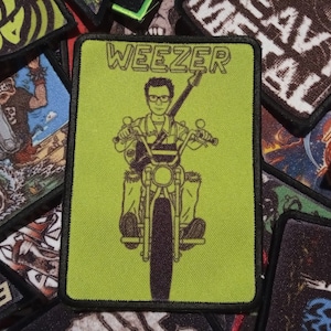 Könnte beinhalten: Ein grüner und schwarzer gestickter Aufnäher mit dem Wort "Weezer" in weißer Schrift. Der Aufnäher zeigt ein Cartoon-Bild einer Person, die ein Motorrad fährt.