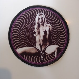 Könnte beinhalten: Ein runder, schwarz-lilafarbener Patch mit einem wirbelnden Muster. In der Mitte des Patches befindet sich ein Schwarzweißbild einer Frau mit Sternen auf der Brust.