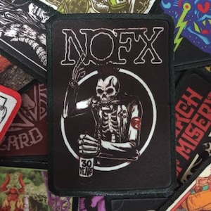 Può includere: Toppa ricamata in bianco e nero con il nome della band "NOFX" in lettere bianche. Uno scheletro con una giacca di pelle e che tiene una birra si trova al centro della toppa. Il testo "30 Years" è sotto lo scheletro.