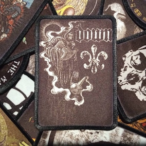 Peut inclure: Un écusson rectangulaire avec un fond marron foncé et une bordure noire. L'écusson présente une illustration blanche d'une figure vêtue d'une robe, une fleur de lys et le mot "IMMORTAL" en blanc. D'autres écussons avec des motifs similaires sont en arrière-plan.