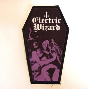 Op de afbeelding: Zwarte en paarse geborduurde patch in de vorm van een kist met de tekst "Electric Wizard" en een kruis boven de tekst. De patch toont een vrouw en een skelet.