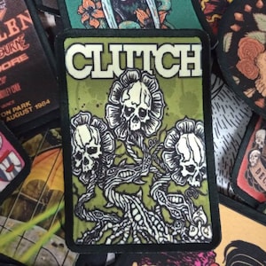 Könnte beinhalten: Ein grüner und schwarzer gestickter Patch mit dem Wort "CLUTCH" in weißen Buchstaben. Der Patch zeigt drei weiße Schädel mit schwarzen Augen und Mündern, jeder mit einem blumenartigen Design. Die Schädel sind durch ein schwarzes und weißes Rankendesign verbunden.