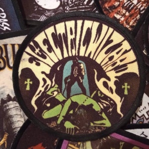 May include: A round, black and white patch with the text "Electric Witch" in a stylised font. The patch features a green and black illustration of a figure with a cross on their chest.