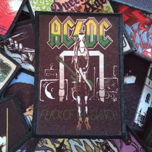 May include: A black and green AC/DC band patch with the band's name in yellow and green letters. The patch features a figure holding a microphone and the words "Flick of the Switch" in white.