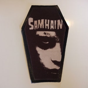 Op de afbeelding: Een geborduurde zwarte en witte patch in de vorm van een kist met het woord "SAMHAIN" in witte letters. De patch heeft een zwart-witte afbeelding van een gezicht.