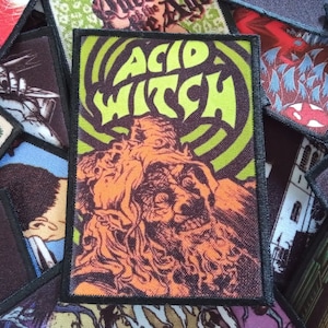 May include: A black and green embroidered patch with the text "Acid Witch" in a green font. The patch features a colourful illustration of a creature with a large head and a long beard.
