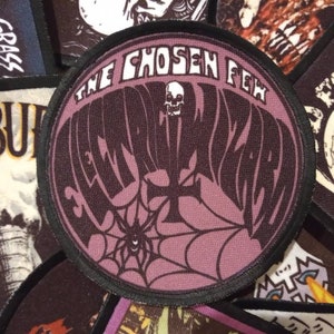 May include: A round, purple and black embroidered patch with the text "The Chosen Few" and "Elemental Wizard" in a stylised font. A spider web with a spider is in the centre of the patch.