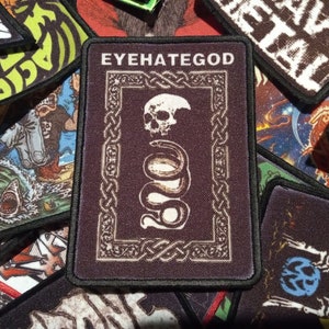 Könnte beinhalten: Ein rechteckiger Aufnäher mit dem Text "EYEHATEGOD" oben, der einen Schädel und ein gewickeltes Schlangendesign zeigt. Der Aufnäher hat einen dunkelbraunen Hintergrund mit schwarzem Rand und einem Rahmen im keltischen Knotenstil. Andere Aufnäher mit verschiedenen Designs befinden sich im Hintergrund.