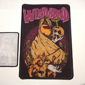May include: A black embroidered patch with a purple and white "WINDHAND" text. The patch features a yellow hooded figure holding a skull with a mushroom growing on top. The figure is surrounded by orange lilies and green foliage.