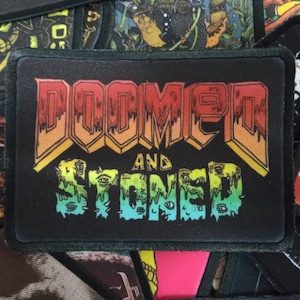 May include: A black patch with a rainbow gradient design. The text "DOOMED AND STONED" is written in a bold, stylized font.