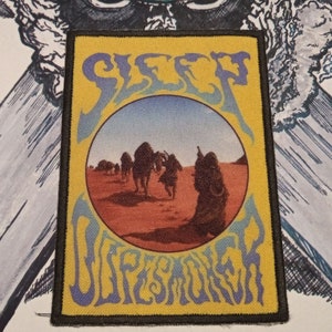 May include: A yellow and blue woven patch with the band name "SLEEP" in a psychedelic font and the album title "Dopesmoker" in a similar font. The patch features a circular image of a desert scene with camels walking across the sand.