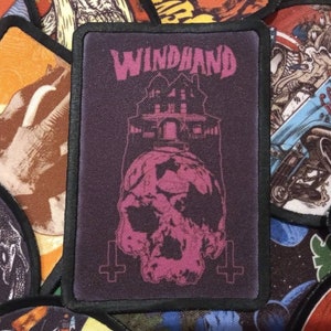 May include: A black and pink embroidered patch with the text "WINDBAND" in white lettering. The patch features a pink skull with a house on top of it.