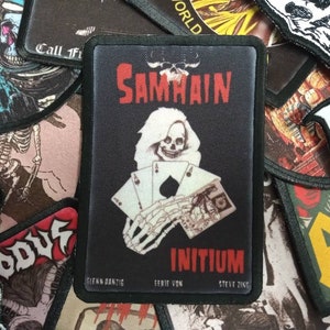 May include: Black and white patch with a skeleton holding playing cards. The text "SAMHAIN" is at the top of the patch and "INITIUM" is at the bottom. The text "GLENN DANTZIG" is in the bottom left corner and "STEVE ZING" is in the bottom right corner.