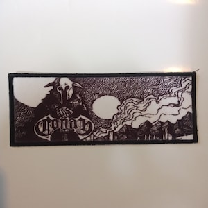 May include: Black and white embroidered patch featuring a hooded figure and the word "Crowning" in a stylised font. The figure is standing in front of a dark, smoky background with a large, white moon.