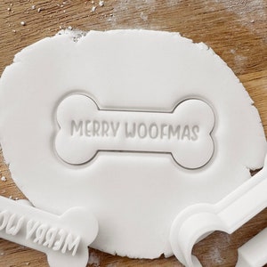 May include: A white bone-shaped cookie cutter with the text "MERRY WOOFMAS" imprinted on white dough. Other white cookie cutters are visible. The scene is set on a wooden surface, suggesting a festive baking theme.