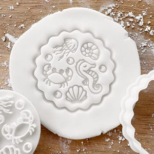 May include: A white, circular cookie cutter with a scalloped edge, featuring embossed sea creatures: a jellyfish, seahorse, crab, seashell, and a spiral shell. The cutter is pressed into white dough, with a second cutter visible.