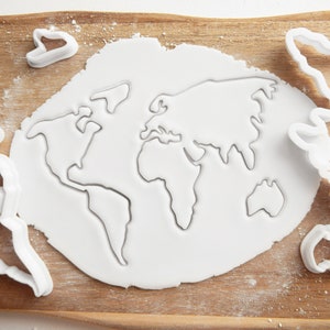 May include: A sheet of white fondant cookie dough with a world map cookie cutter pressed into it. The cookie cutter is made of white plastic and is visible around the edges of the dough.