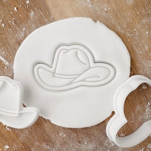 May include: A white cowboy hat-shaped cookie cutter pressed into white dough, with two other cutters nearby. The scene is set on a wooden surface dusted with flour, suggesting a baking or crafting theme.