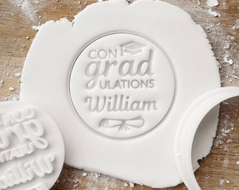 May include: White fondant with a round cookie cutter imprint that reads "Congrats grad William" with a graduation cap and diploma graphic.