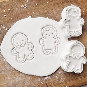 May include: White plastic gingerbread person cookie cutters and a circle of white dough on a wooden surface. Two cutters are shaped like gingerbread people with bow ties, and two are shaped like gingerbread people with bows.