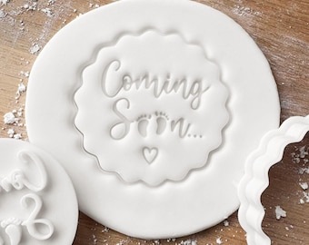 May include: White plastic cookie cutter with a scalloped edge. The cutter is shaped like a circle with the words "Coming Soon..." and a pair of baby footprints inside.