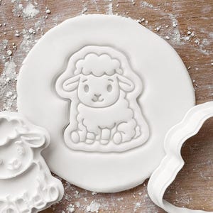 May include: A white sheep-shaped cookie cutter pressed into a round of white dough. The sheep has a fluffy body, small ears, and a smiling face. A second sheep-shaped cutter and a wavy-edged cutter are also visible.