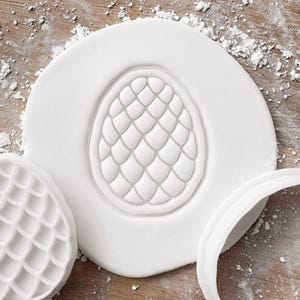May include: A white, egg-shaped cookie cutter pressed into a circle of white dough. The cutter has a textured, scale-like pattern. A separate, similar cutter and a curved edge are also visible on a wooden surface dusted with flour.