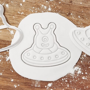 May include: White alien-shaped cookie cutter on a white dough circle. The cutter features a one-eyed alien in a spaceship design. The background is a wooden surface with flour.