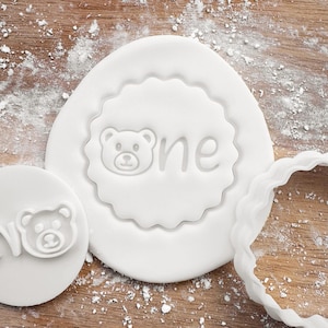 May include: White fondant with a bear and the word "one" imprinted on it. The fondant is on a wooden surface with white powder.