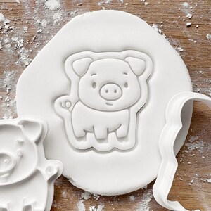 May include: A white pig-shaped cookie cutter pressed into white dough. The cutter has a detailed outline of a pig with a smiling face, small ears, and a curly tail. The background is a wooden surface with flour.