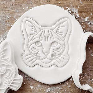 May include: A white cat-shaped cookie cutter and stamp set. The stamp features a detailed cat face design, while the cutter has a curved edge. The set is on a wooden surface with flour dusting.