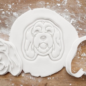May include: A white plastic cookie cutter in the shape of a dog's head. The cutter is on a white surface with a light brown background.