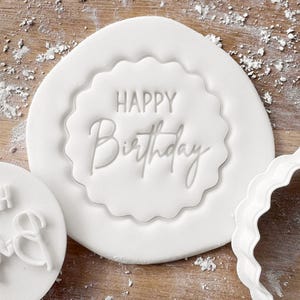 May include: A white fondant stamp with the words "HAPPY Birthday" in a script font. The stamp has a scalloped edge and is surrounded by a white fondant base. Additional white fondant pieces are visible.