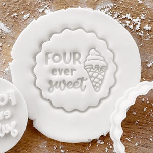 May include: White cookie cutter with a scalloped edge. The cutter is shaped like a circle with the words "FOUR ever sweet" and a cartoon ice cream cone inside.