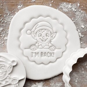 May include: White cookie cutter featuring an elf design and the text "I'M BACK!". The elf is wearing a hat and smiling. The cutter is on a white surface with a scalloped edge, with other cutters visible.