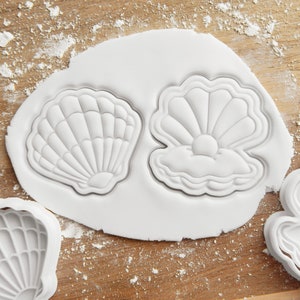 May include: Two white plastic cookie cutters shaped like seashells. One seashell has a pearl inside. The cutters are on a white surface covered in icing sugar.