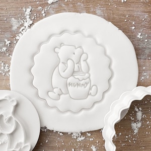 May include: A white cookie cutter featuring Winnie the Pooh. The design shows Pooh Bear with a honey pot, the word "Hunny" is visible. The cutter is round with a scalloped edge. It is on a wooden surface.