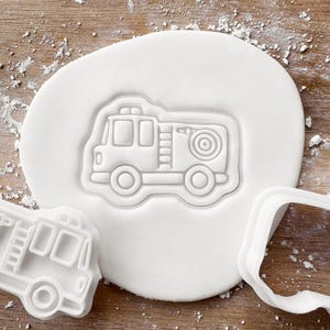 May include: A white, fire engine-shaped cookie cutter pressed into a circle of white dough. The cutter is next to a cut-out shape of the same fire engine design. The background is a light brown wooden surface.