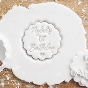 May include: A white fondant surface with a clear plastic cookie cutter imprinted on it. The cutter is shaped like a circle with a scalloped edge and says "Nylah's 1st Birthday" in a cursive font.