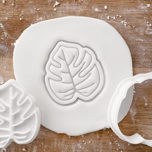 May include: A white fondant leaf cookie cutter with a detailed monstera leaf design. The cutter is shown pressed into white fondant.