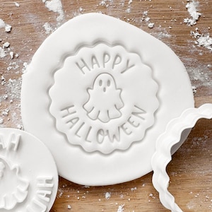 May include: White Halloween cookie stamp with a scalloped edge. The stamp features a ghost illustration and the words "HAPPY HALLOWEEN" in a circular design. Additional cookie stamp visible.