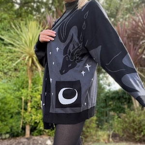 Peut inclure: Cardigan noir et gris avec un motif de dragon, des étoiles et une poche en forme de croissant de lune. Le pull à manches longues a une fermeture boutonnée et est porté sur un col roulé.