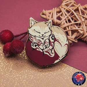 Könnte beinhalten: Eine goldfarbene Emaille-Anstecknadel mit einer weißen Katze, die sich in einer burgunderfarbenen Herzform zusammengerollt hat. Die Katze hat geschlossene Augen und eine kleine Pfote ist sichtbar. Die Anstecknadel hat einen goldfarbenen Rand und ein schwarz-weißes Logo mit dem Text "Chocolate Raisin Fury" in einem Kreis.