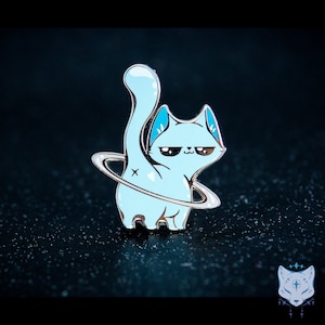 Könnte beinhalten: Eine blaue Emaille-Anstecknadel mit einem blauen Ring um den Schwanz einer Katze. Die Katze hat einen mürrischen Gesichtsausdruck und schaut nach rechts. Die Anstecknadel befindet sich auf einem schwarzen Hintergrund.