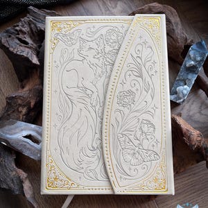 May include: A cream-colored journal with a magnetic flap closure featuring intricate gold foil embossed line art of a fox, butterflies, and flowers. The cover design includes decorative borders and star accents, with a smooth, matte finish.