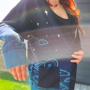 May include: A navy blue cardigan sweater with a celestial design. The sweater features white star and cloud patterns, a dark blue pocket, and light blue trim. The cardigan has a V-neck and button closure.