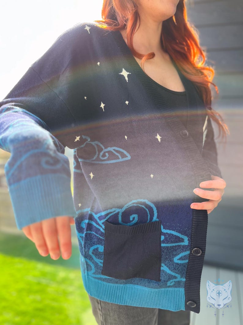 May include: A navy blue cardigan sweater with a celestial design. The sweater features white star and cloud patterns, a dark blue pocket, and light blue trim. The cardigan has a V-neck and button closure.