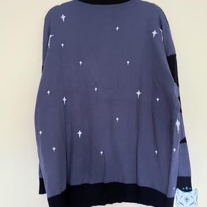 May include: A dark blue sweater with white star patterns. The sweater has a black collar, cuffs, and hem. The back of the sweater is visible, hanging on a purple hanger. The sweater is a knitwear item.